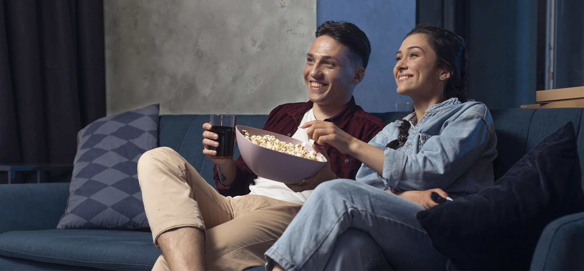 A young couple eating popcorn and watching a movie on their couch